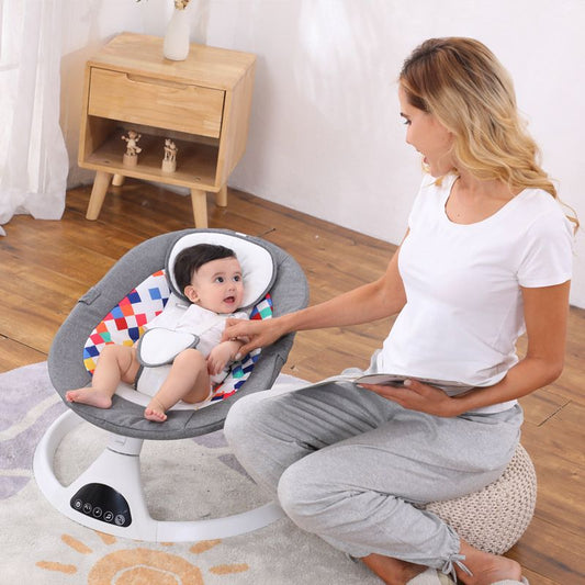 Bébé dans une balancelle électrique grise avec motifs, écoutant une femme lisant à côté, dans une pièce éclairée avec déco simple